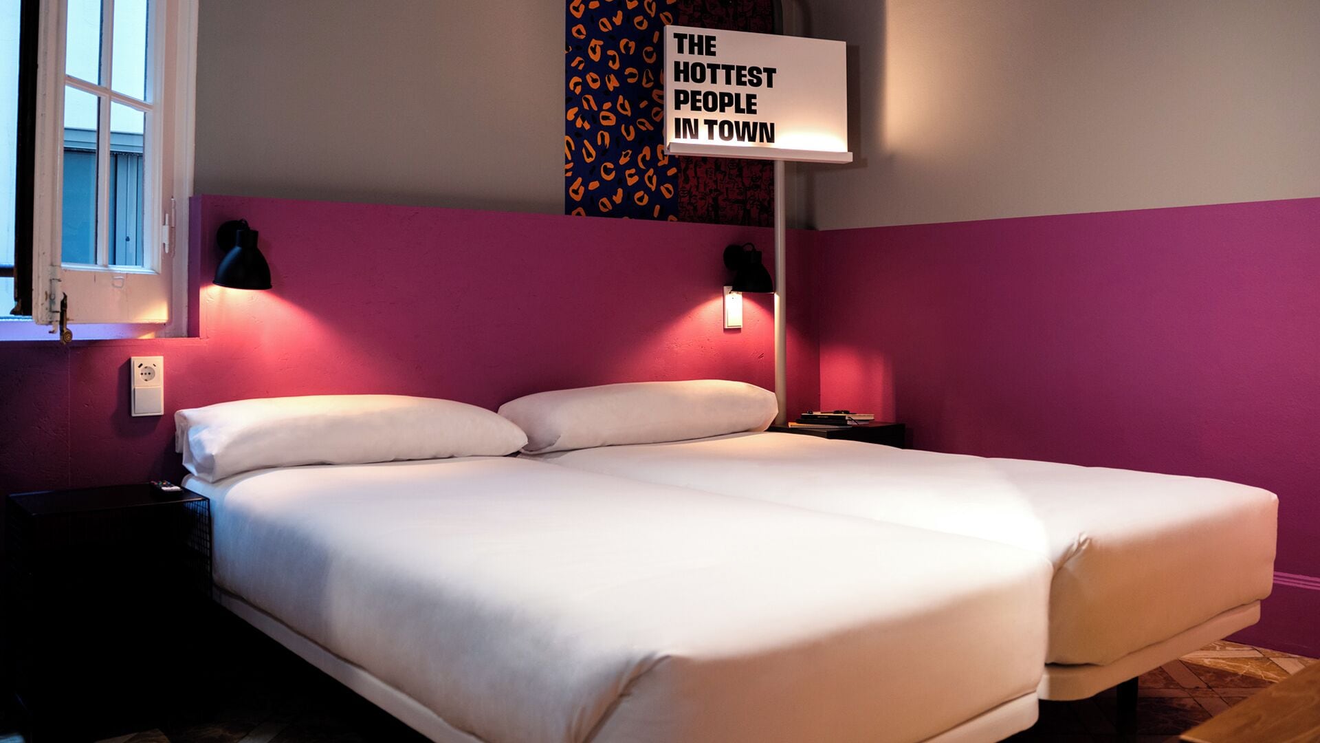 A hotel room with a bed, two bedside tables, a nightstand, a window, and a sign on the wall.