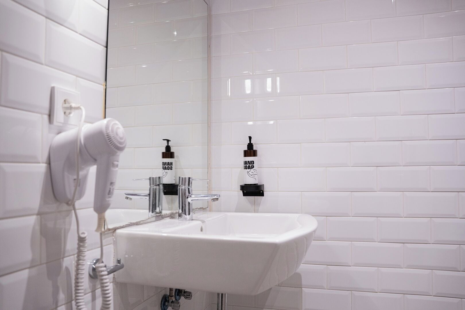 Un baño con azulejos blancos, un lavamanos blanco, un secador de pelo y dos dispensadores de jabón para manos.