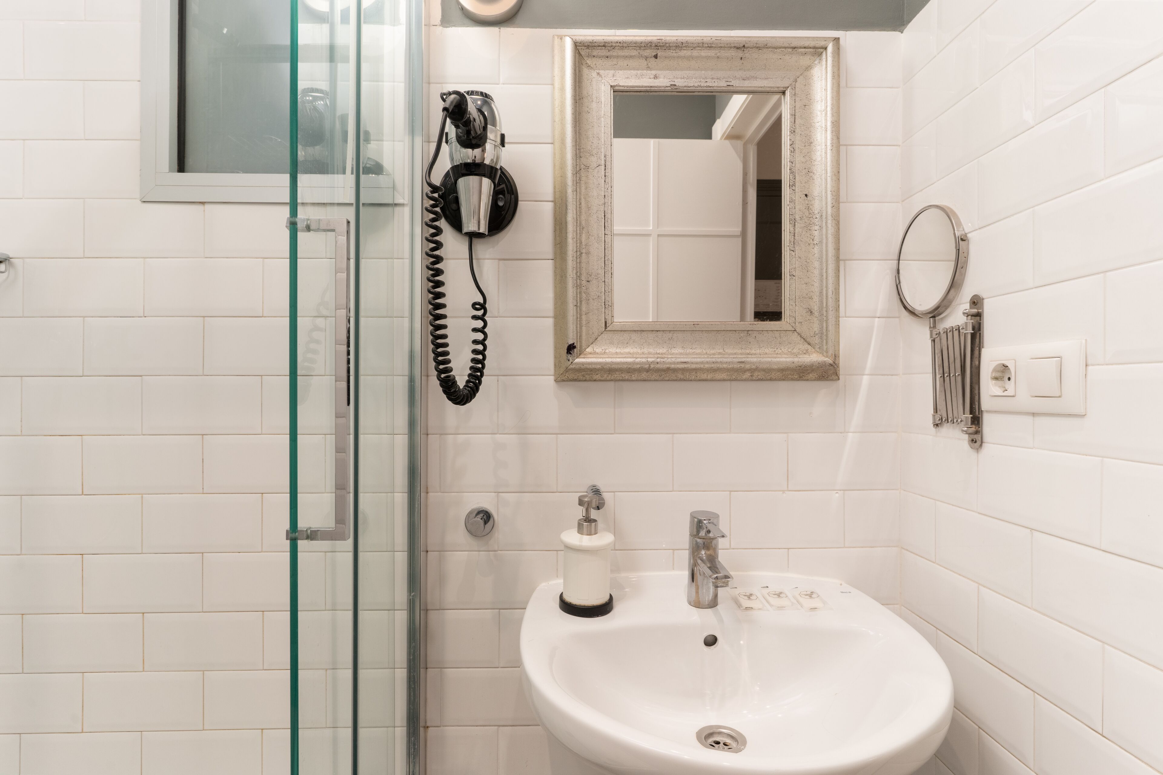 Un baño con cabina de ducha de vidrio, un lavamanos blanco con espejo encima y un teléfono fijado a la pared.