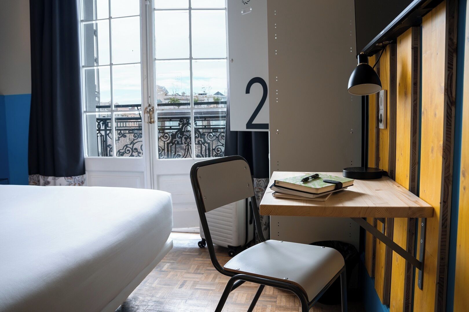 Et hotelværelse med en seng, et skrivebord med stol, en lampe og udsigt over byen gennem et vindue.