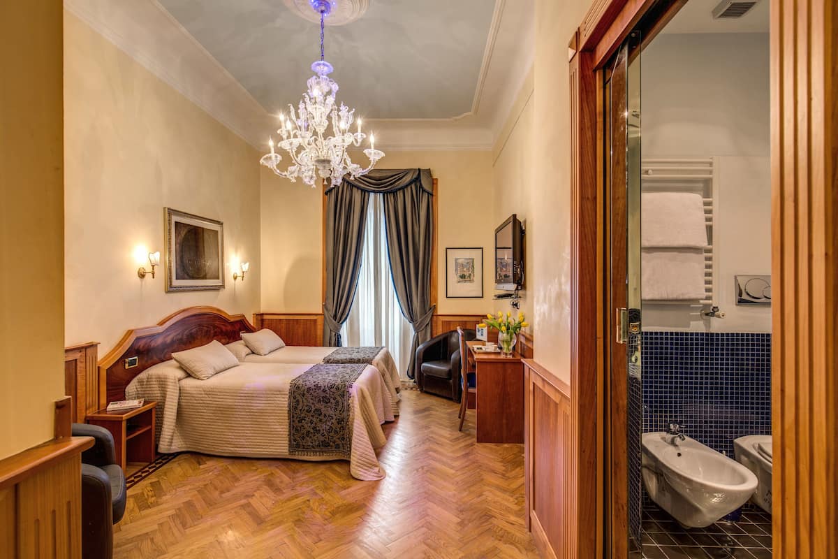 A hotel room with a large bed, a desk, a chair, a chandelier, and a bathroom visible through an open door.