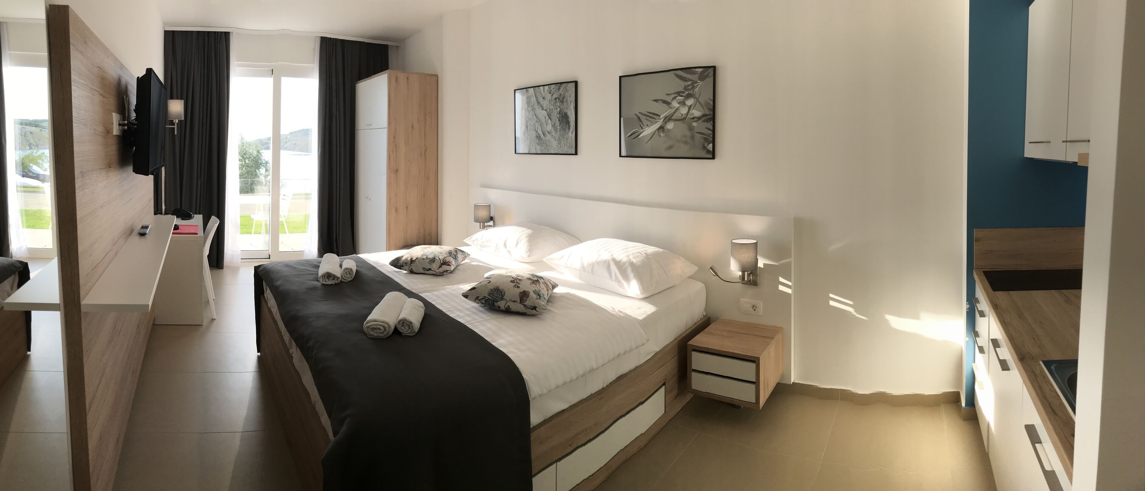 A modern hotel room with a bed, bedside tables, a TV, artwork, and a view of the outdoors.
