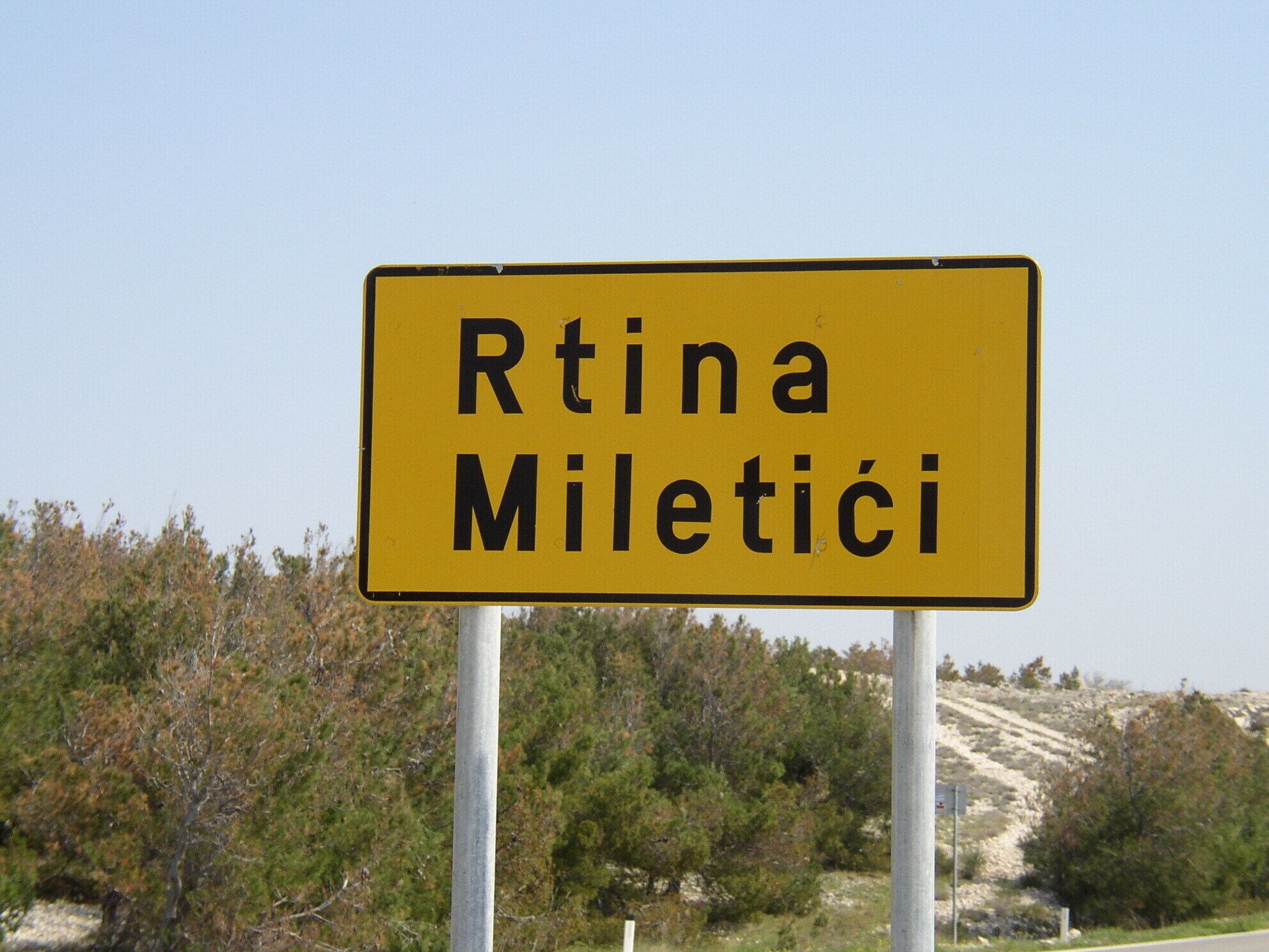Una señal de tráfico amarilla con el texto "Rtina Miletići" sobre un fondo de árboles y un cielo despejado.