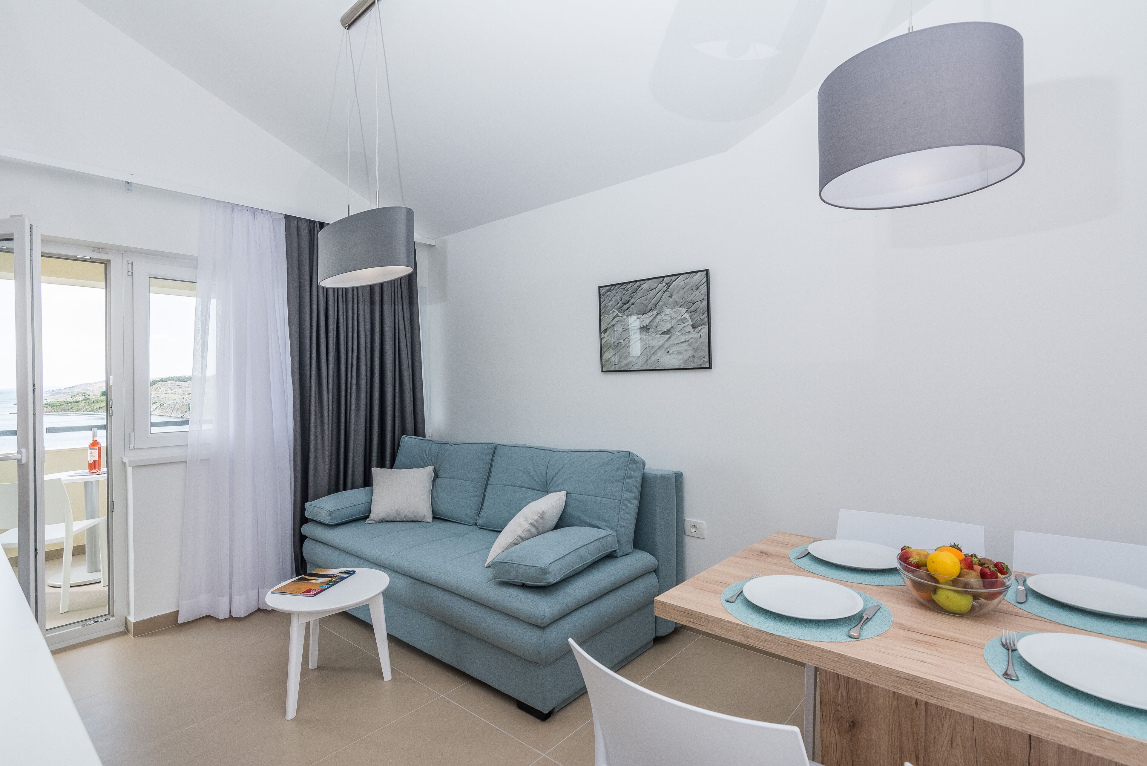 Un salón moderno con un sofá, una mesa de comedor con platos y un cuenco de fruta, y vistas a través de un ventanal.