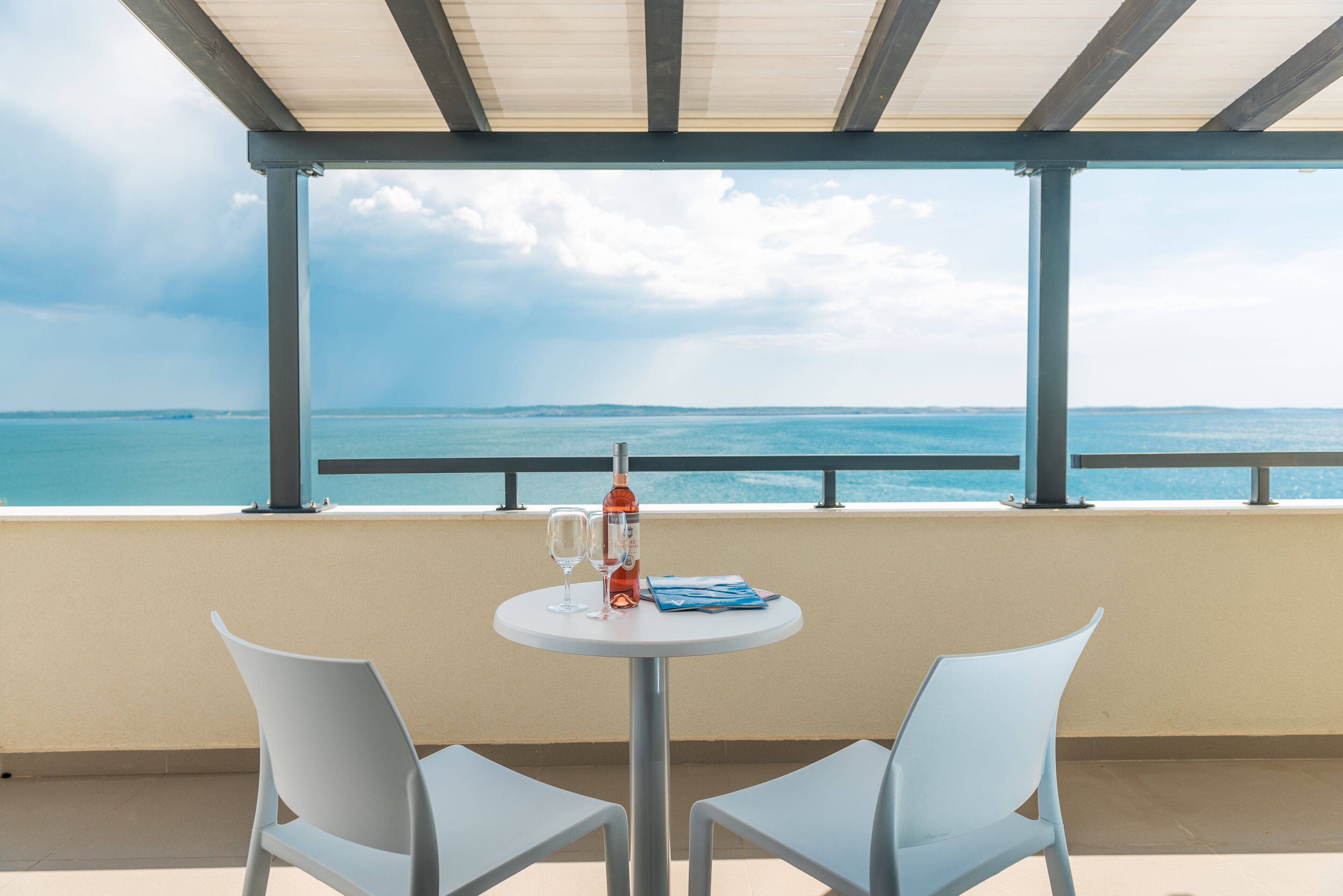 A table with two chairs and a bottle of wine on a balcony overlooking the sea.