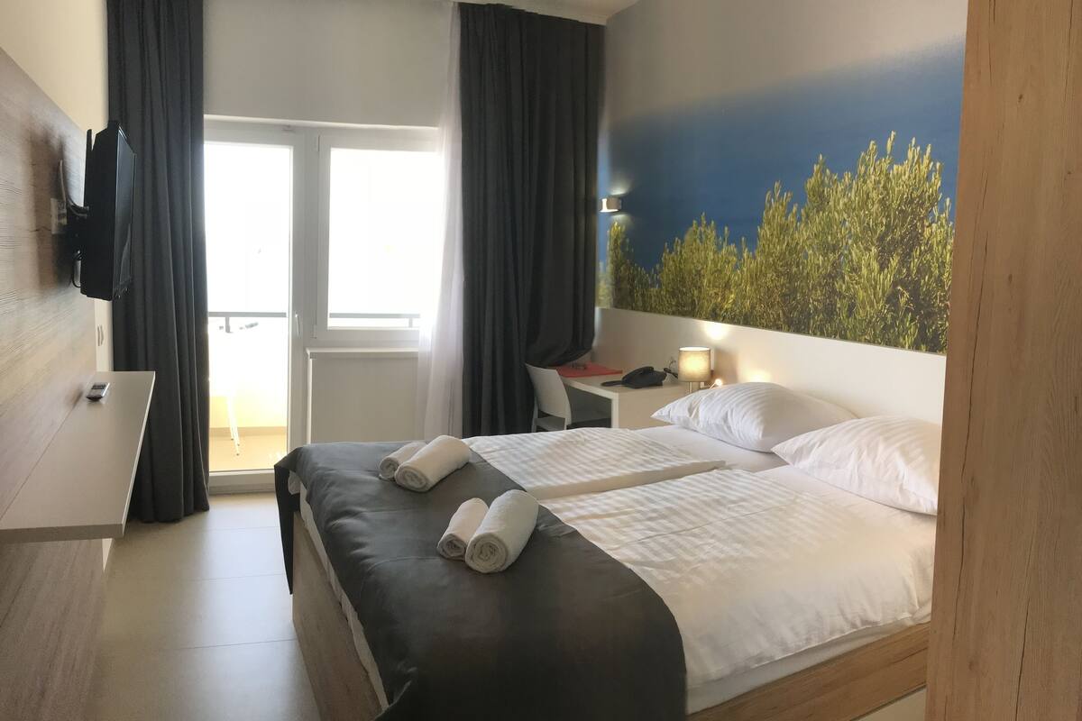 Ein Hotelzimmer mit Bett, Schreibtisch, Stuhl, Fernseher und Blick auf Grünflächen durch das Fenster.