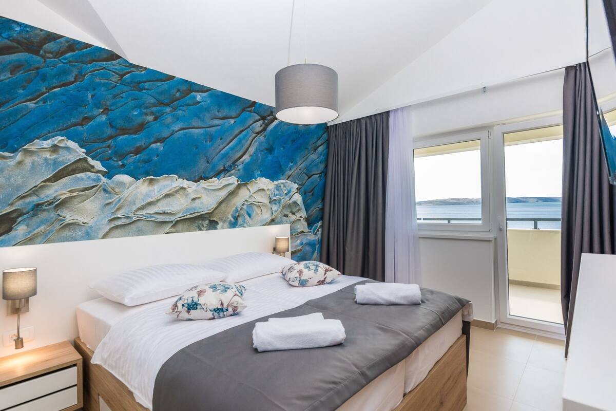 Ein Schlafzimmer mit einem großen Bett, einer Kopfteil mit einem blauen, abstrakten Wandbild, einem Nachttisch mit Lampe und Blick auf das Meer durch ein Fenster.