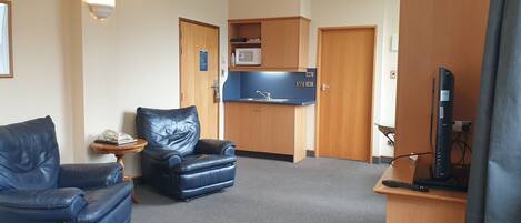 A compact hotel room with a leather sofa, a small armchair, a kitchenette with a microwave, and a television.