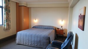Una habitación de hotel con una cama, una silla, una mesita de noche con una lámpara y un cuadro en la pared.