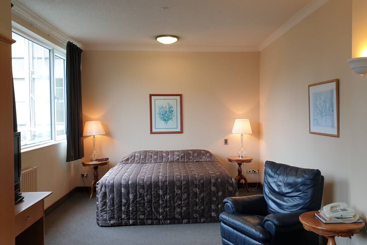 A hotel room with a bed, a chair, two bedside tables with lamps, and two framed pictures on the walls.