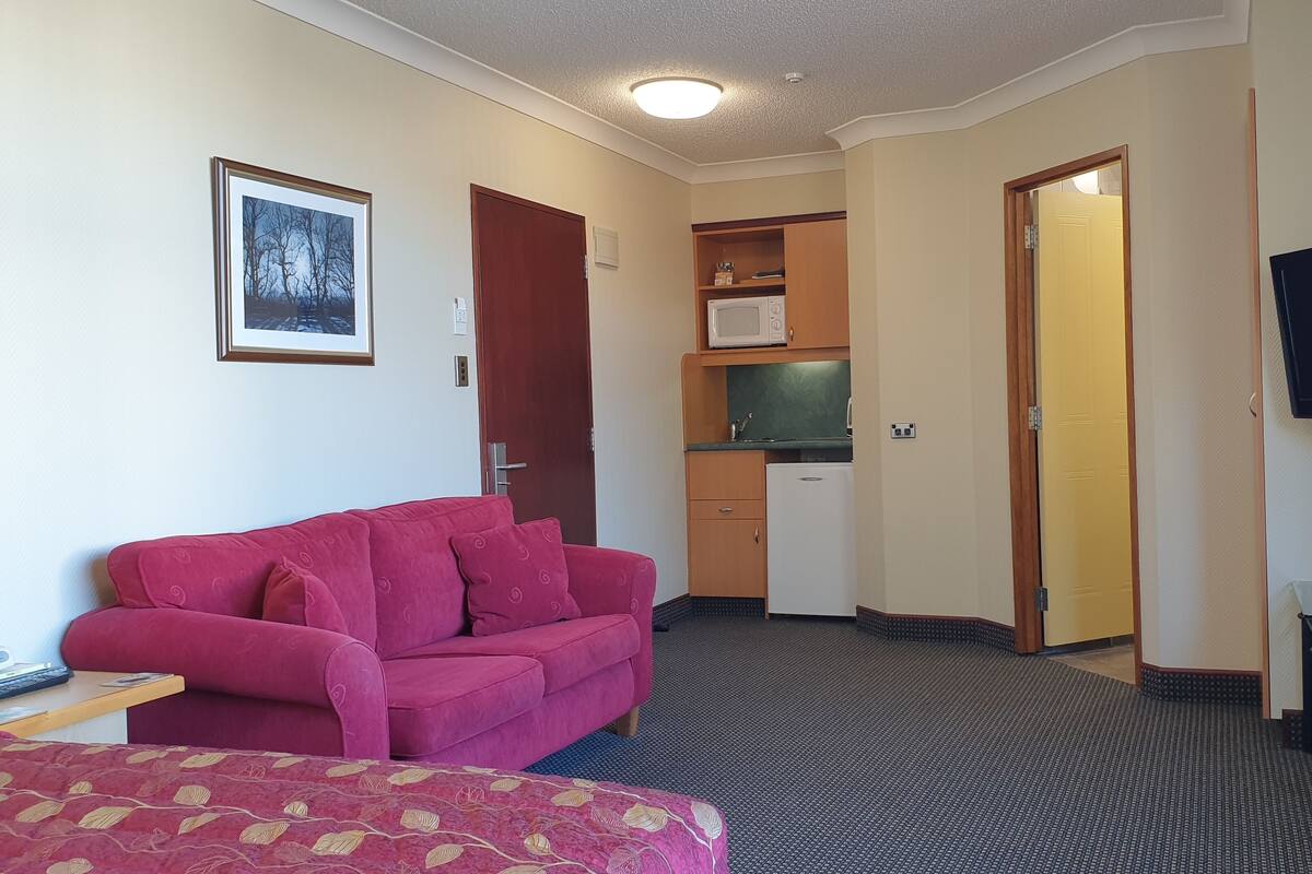 A compact hotel room with a red sofa, a small kitchenette, a TV, and a bed with a patterned cover.
