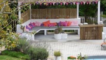 Relax in a serene outdoor space surrounded by lush greenery, featuring a covered seating area with a bench and cushions, a small table, and a pool.