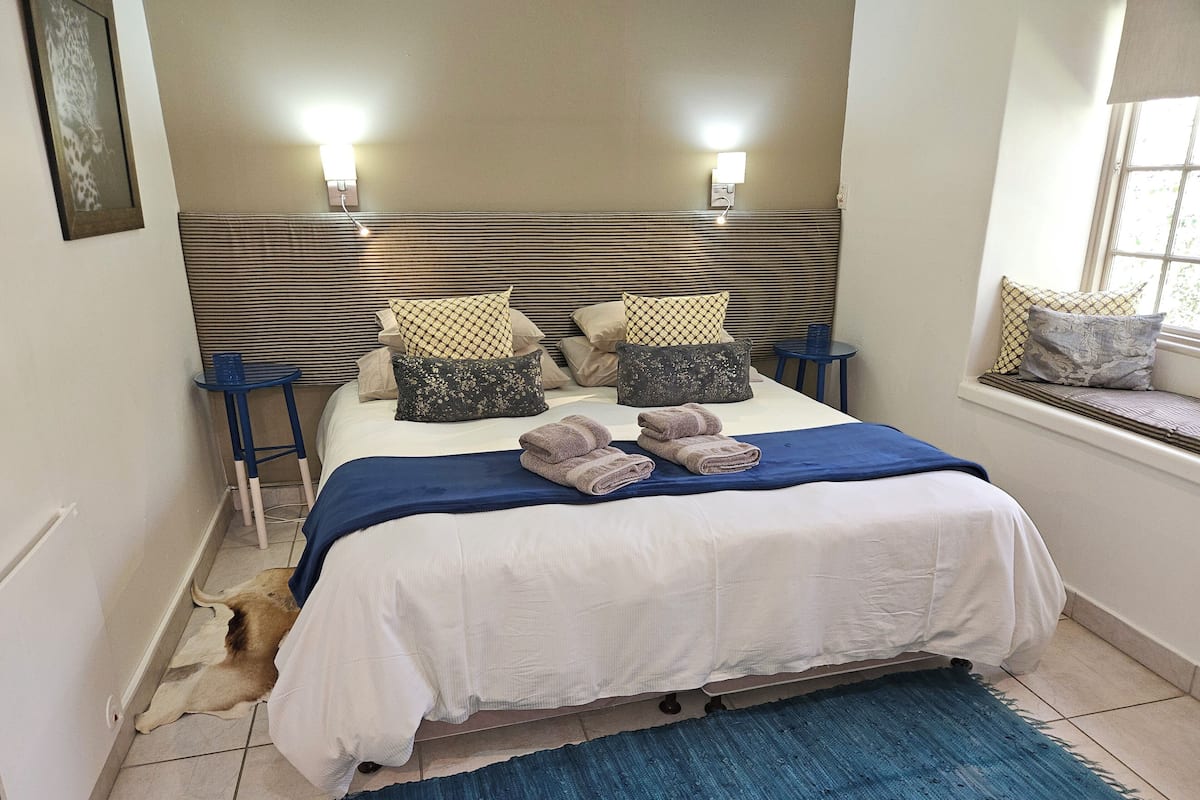 Una cama bien hecha con una manta azul, almohadas y toallas dobladas. Hay una mesita azul al lado de la cama, y un rincón con cojines junto a la ventana.