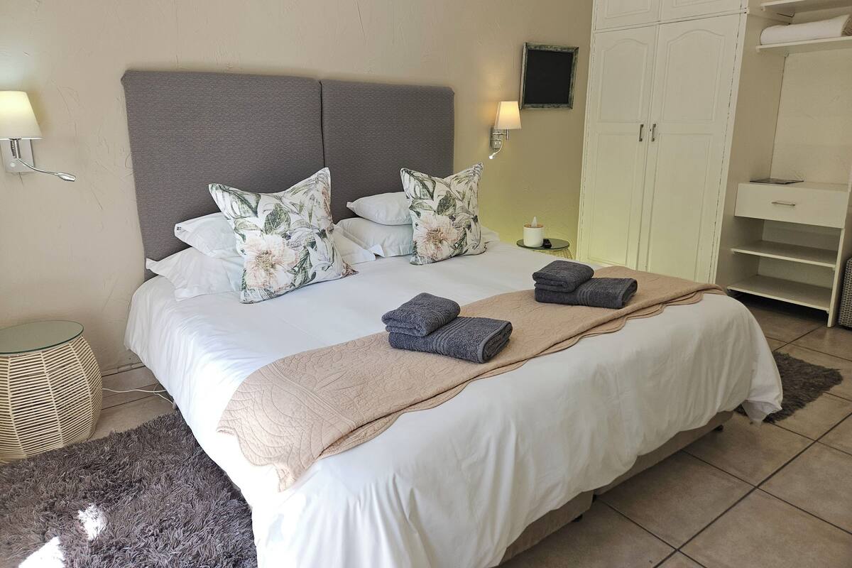 Una cama bien hecha con cabecera gris, ropa de cama blanca y almohadas florales. Hay dos lámparas de noche, una mesita de noche con una vela y un armario con estantes abiertos.