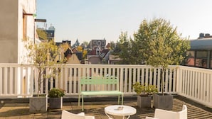 Terraza en la azotea con sillas blancas y una mesa pequeña, rodeada de plantas en macetas y una baranda blanca.