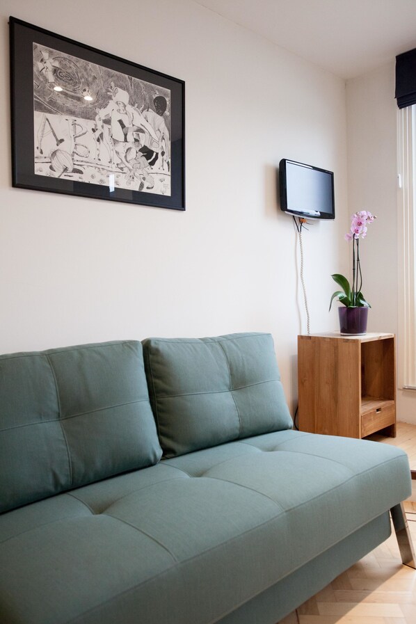 A modern living room with a teal sofa, a framed black and white artwork, a flat-screen TV, and a potted orchid. - Kuwadro Guesthouse Centrum (Amsterdam)