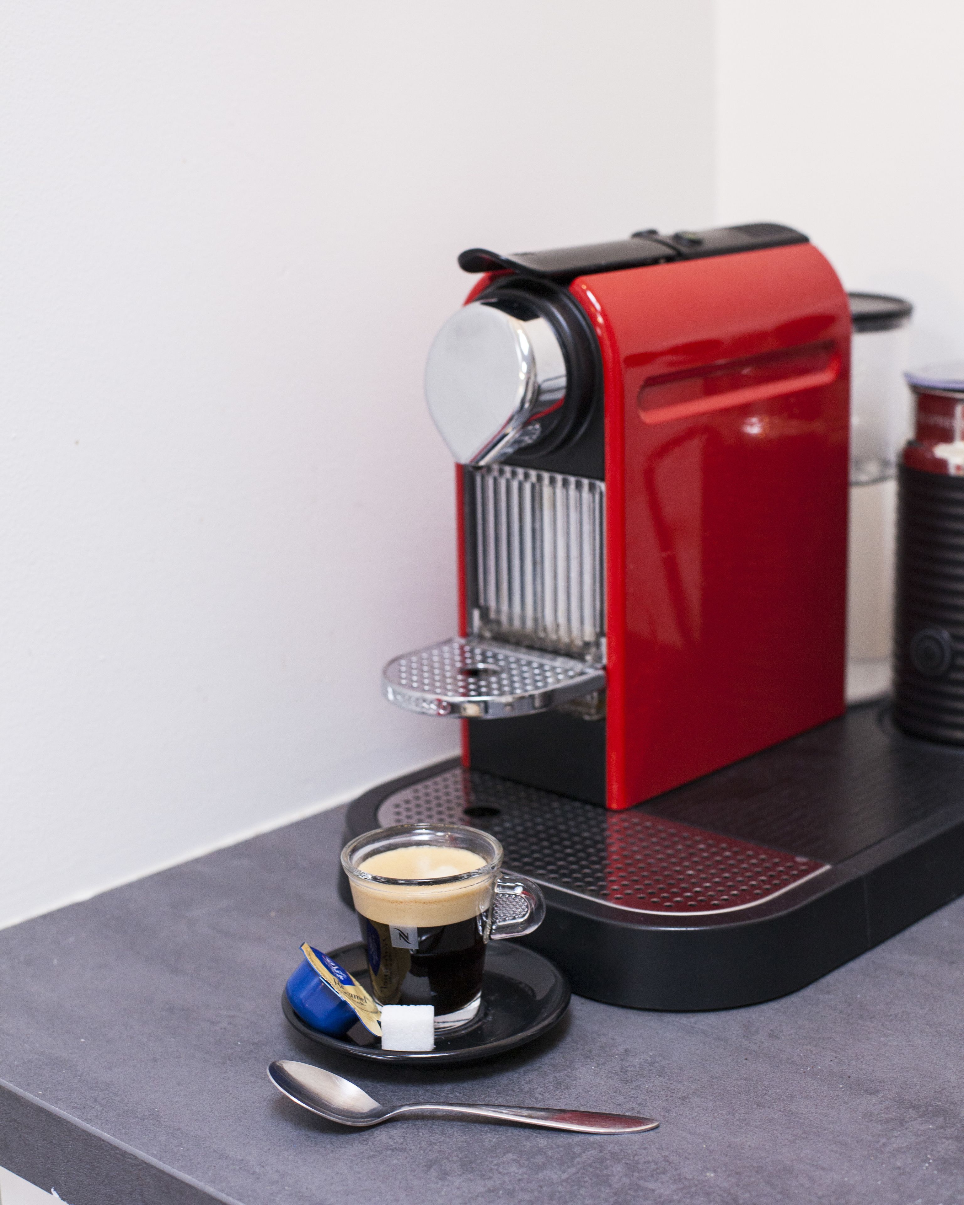 A red espresso machine with a glass cup of coffee on a black tray, a spoon, and a small packet of sugar.