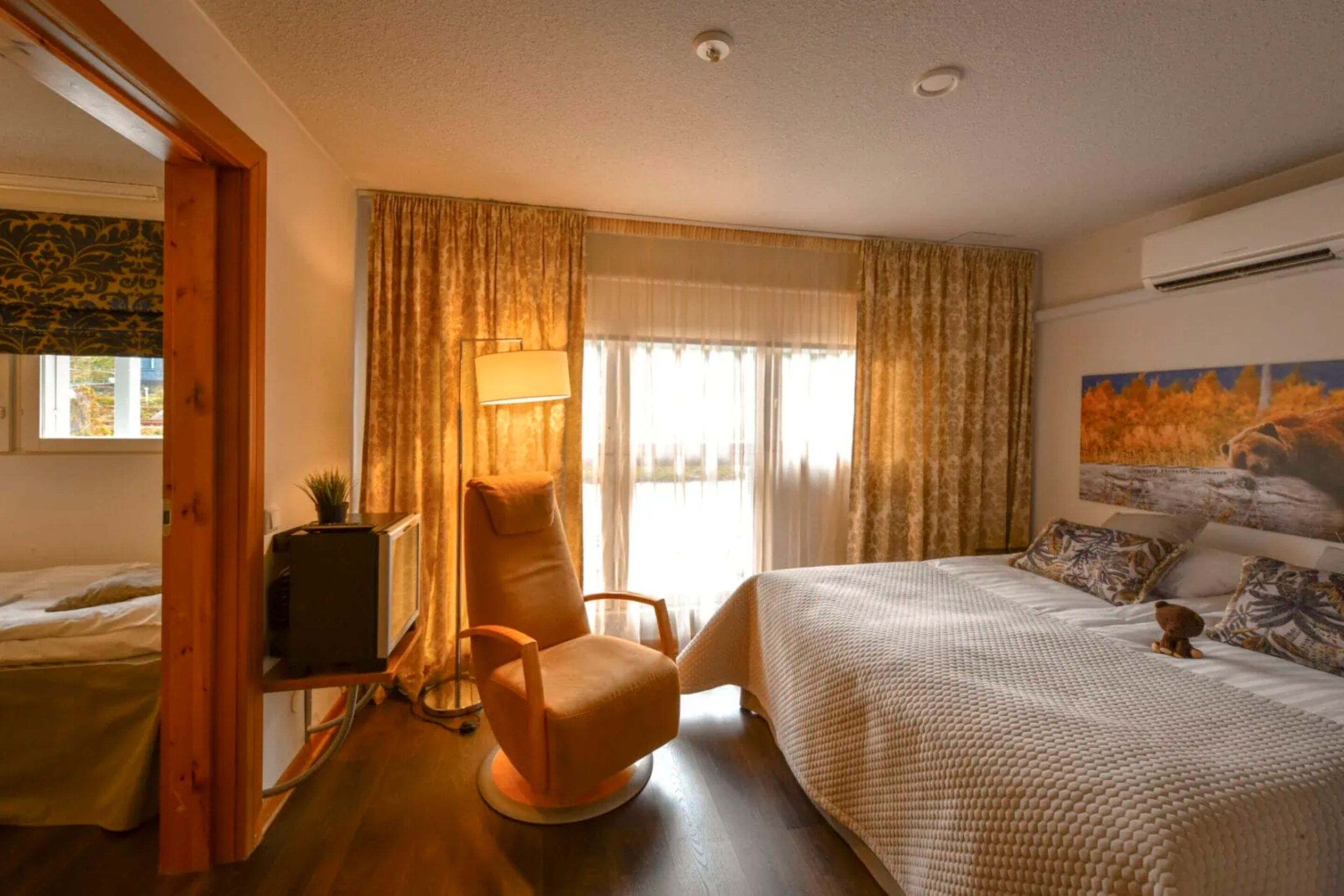 A hotel room with a bed, a chair, a lamp, a painting, and a window with curtains.