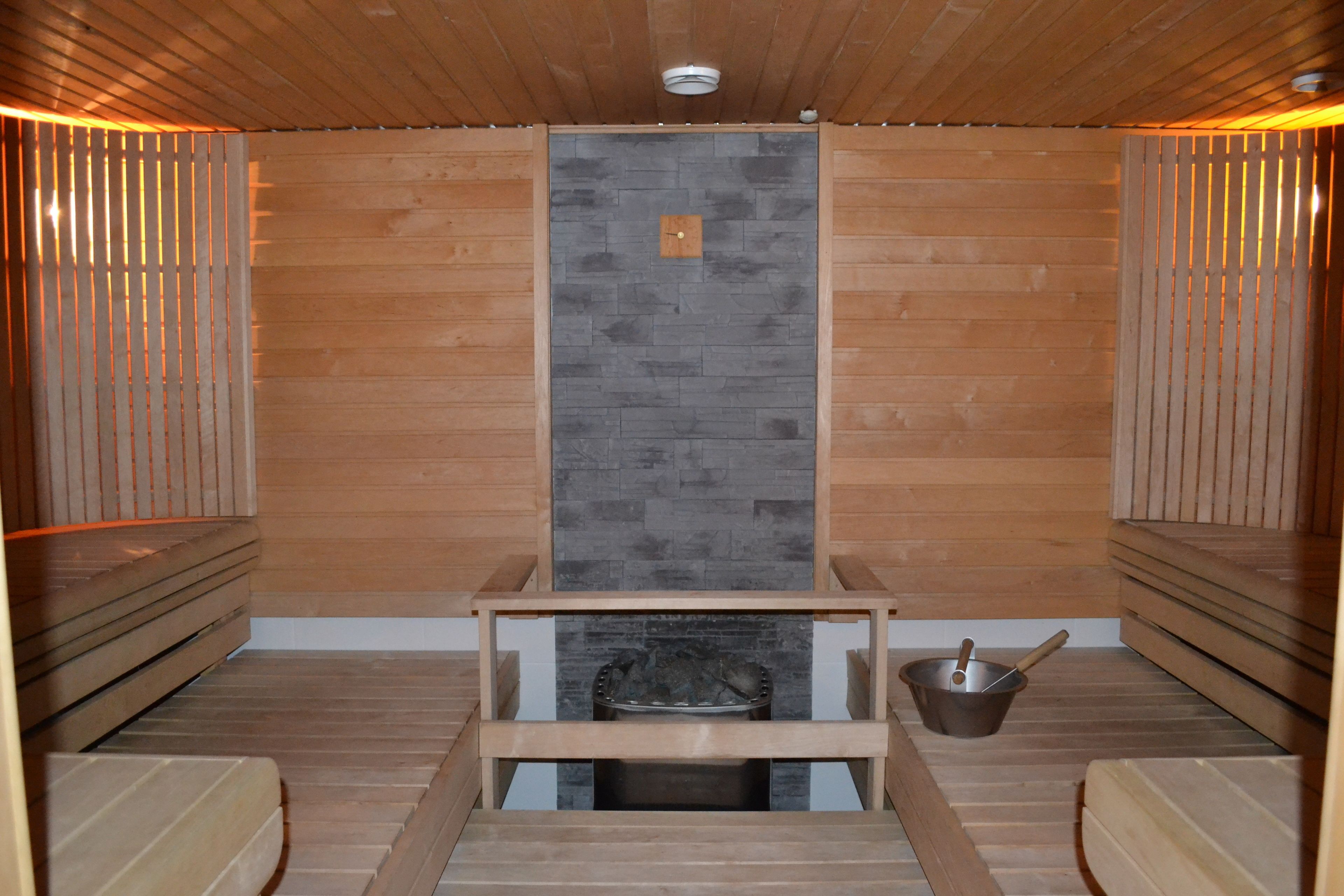 Una sauna con paredes y bancos de madera, una pared central de piedra y un cubo metálico con una cuchara.