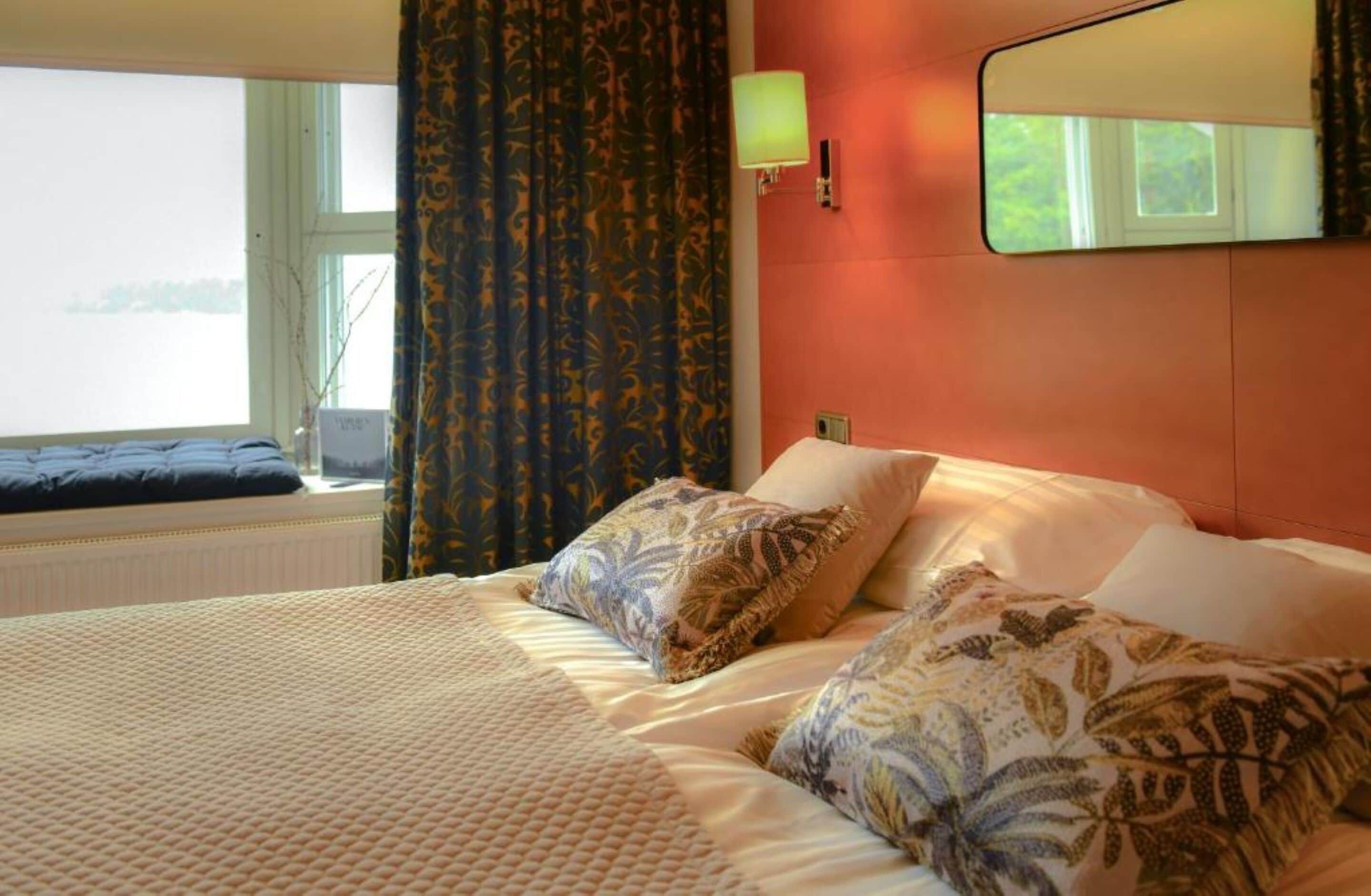 A hotel room with a bed, a window with curtains, a lamp, and a mirror on the wall.