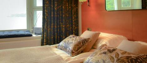 Una habitación de hotel con cama, ventana con cortinas, lámpara y espejo en la pared.