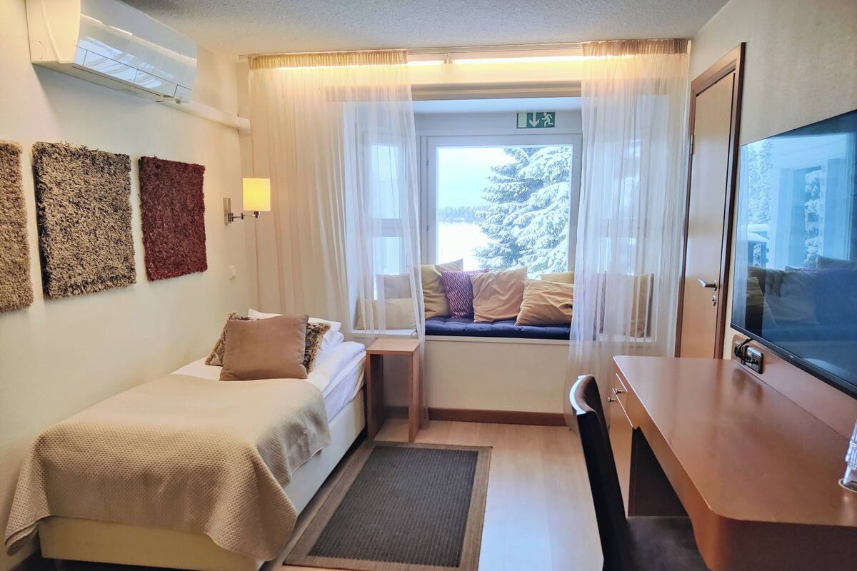 Ein Hotelzimmer mit Bett, Schreibtisch, Stuhl, Fernseher und einem Fenster mit Vorhängen sowie Blick auf schneebedeckte Bäume.