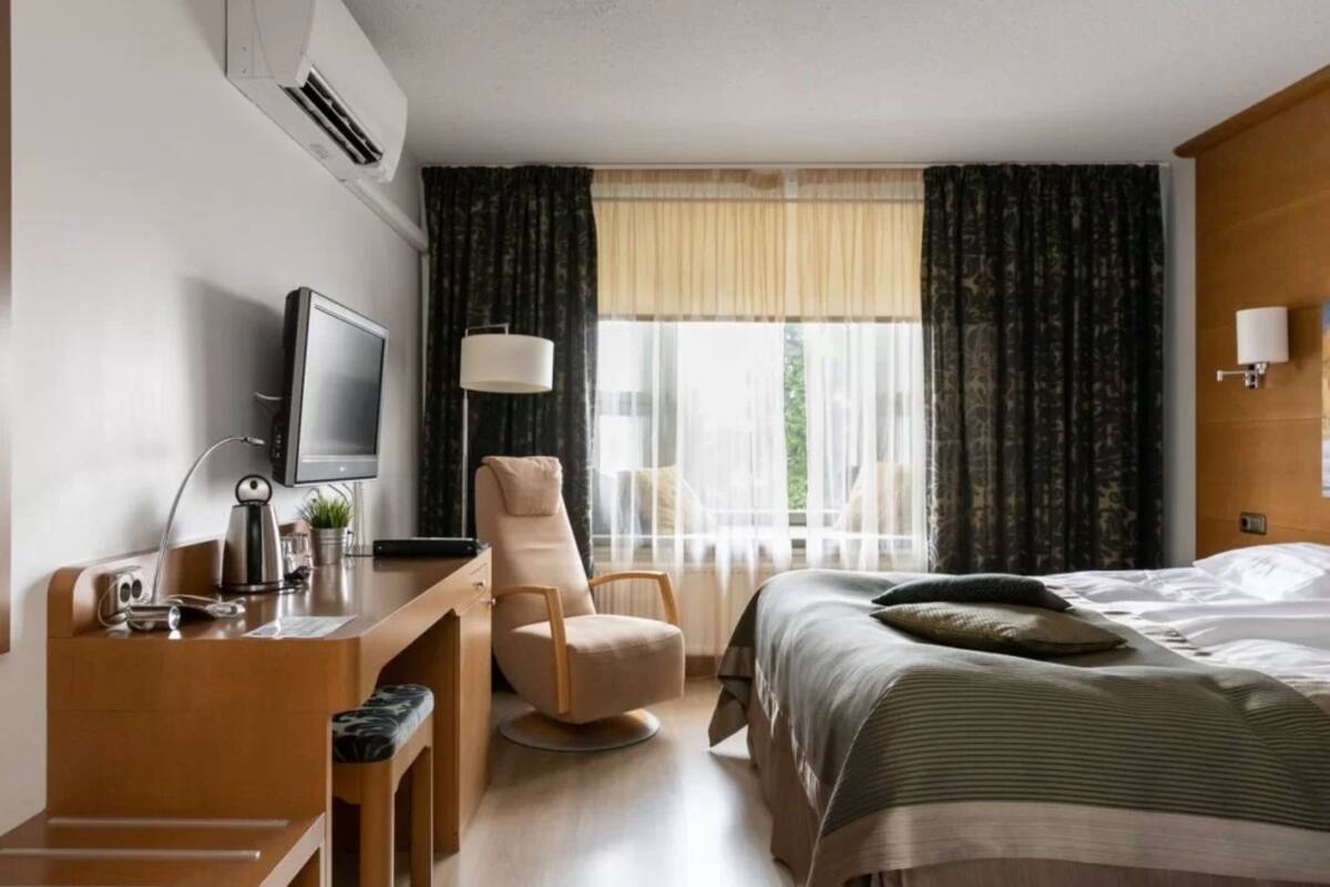 Ein Hotelzimmer mit einem Bett, einem Schreibtisch mit Computer, einem Sessel, einem Fernseher und einem Fenster mit Vorhängen.