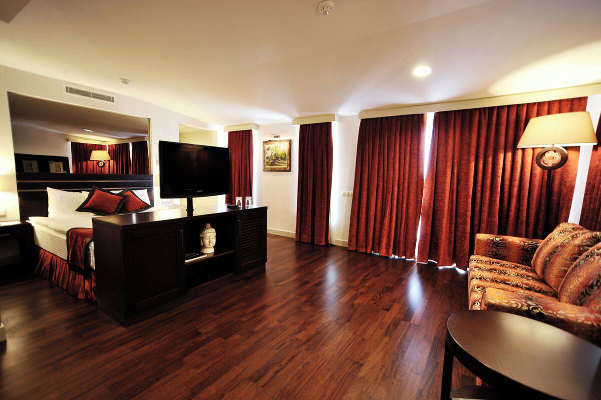 A hotel room with a bed, a TV, a desk, a chair, a sofa, and a large window with curtains.