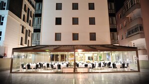 Ein Hotel mit Glasfassade, mehreren Etagen und einem überdachten Außenbereich mit Tischen und Stühlen.