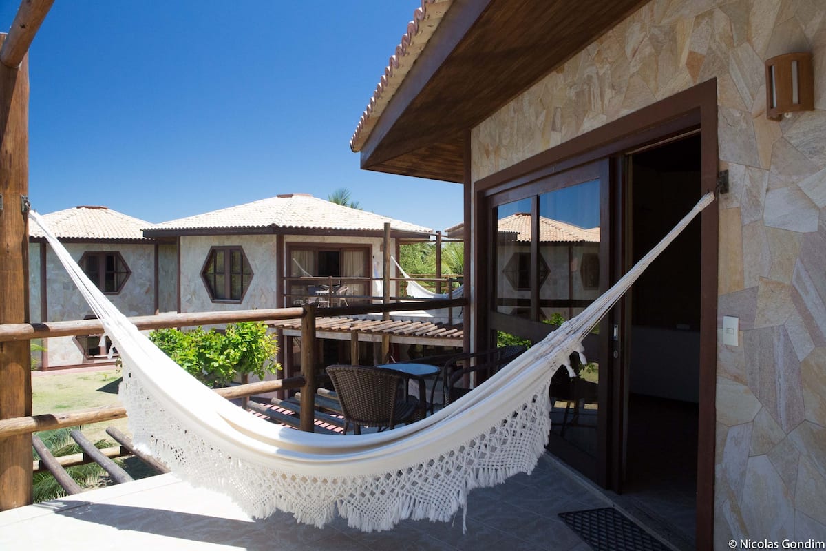 Un hamac sur un balcon, avec vue sur un bâtiment aux murs en pierre et aux toits couverts de tuiles.