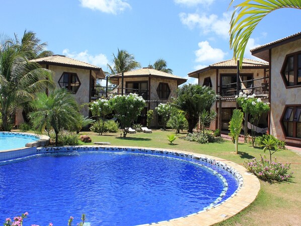 A pool surrounded by a garden with palm trees and a building with a tiled roof. - Morada dos Ventos (Tibau do Sul)
