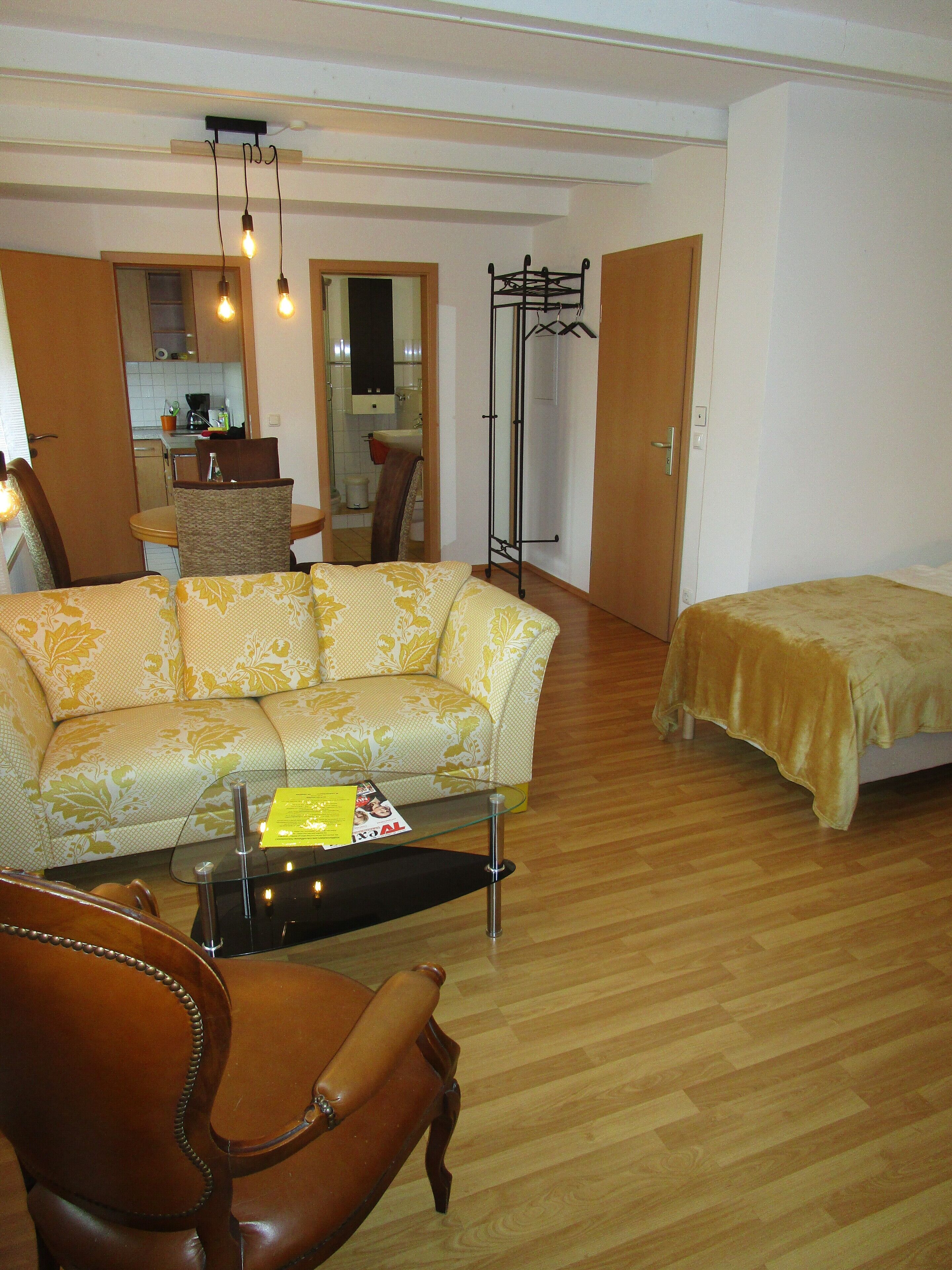 A living room with a sofa, a coffee table, a chair, and a bed. There is a kitchen in the background.