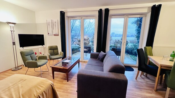 Una sala de estar moderna con un sofá, sillas, una mesa de centro y un televisor. Amplias ventanas ofrecen vistas al exterior.