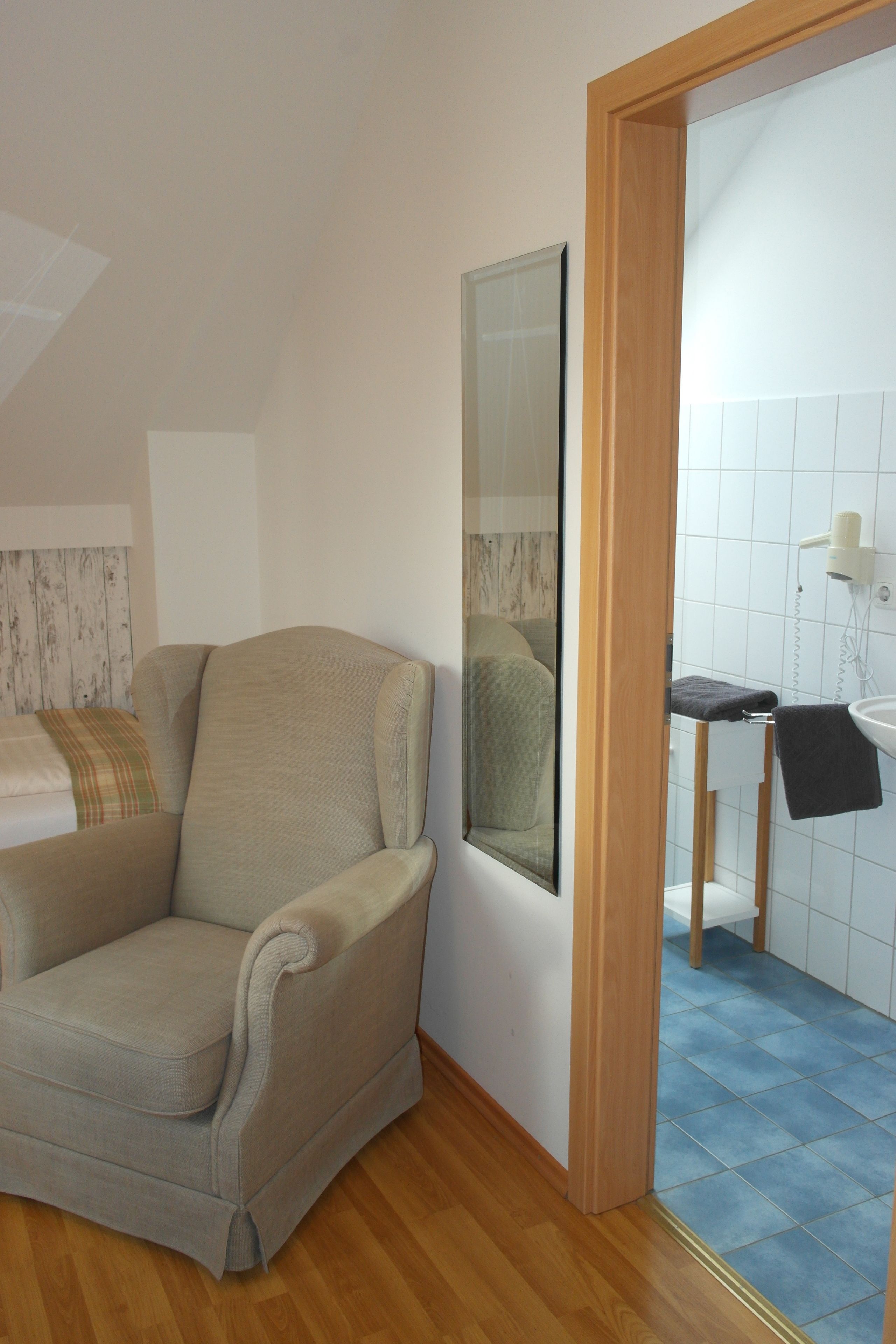 A room with a beige armchair, a wooden door leading to a bathroom with a sink and mirror, and a wooden floor.