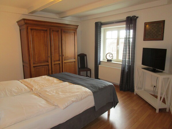Un dormitorio con cama, armario de madera, una silla, ventana con cortinas, radiador y televisor sobre un mueble.