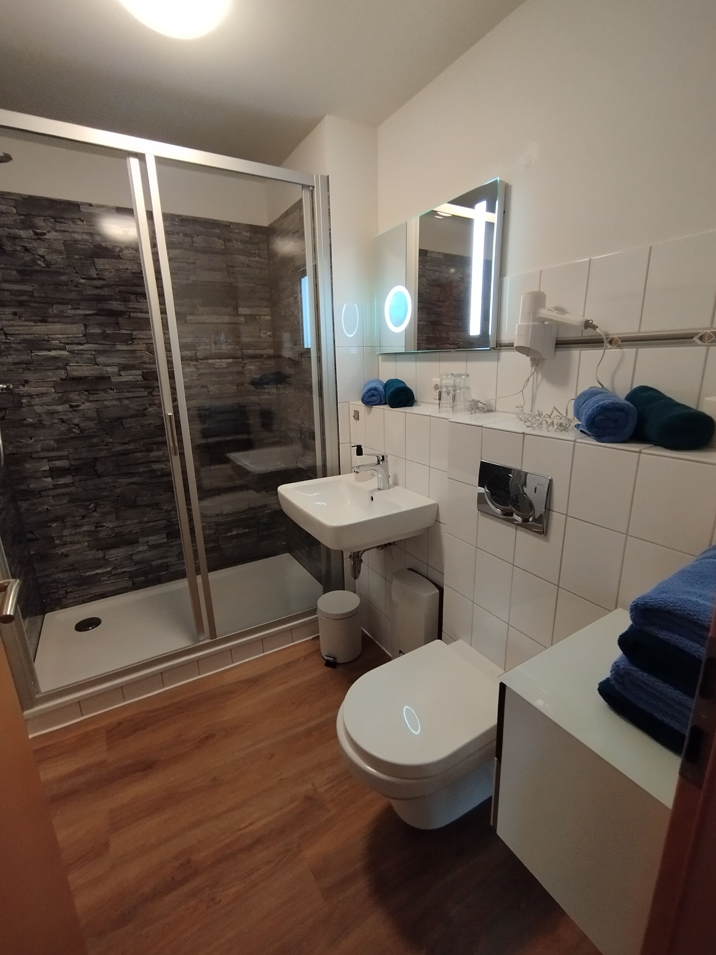 Ein modernes Badezimmer mit Duschabtrennung aus Glas, einem weißen Waschbecken, einer Toilette und einem Waschtisch mit Handtüchern.