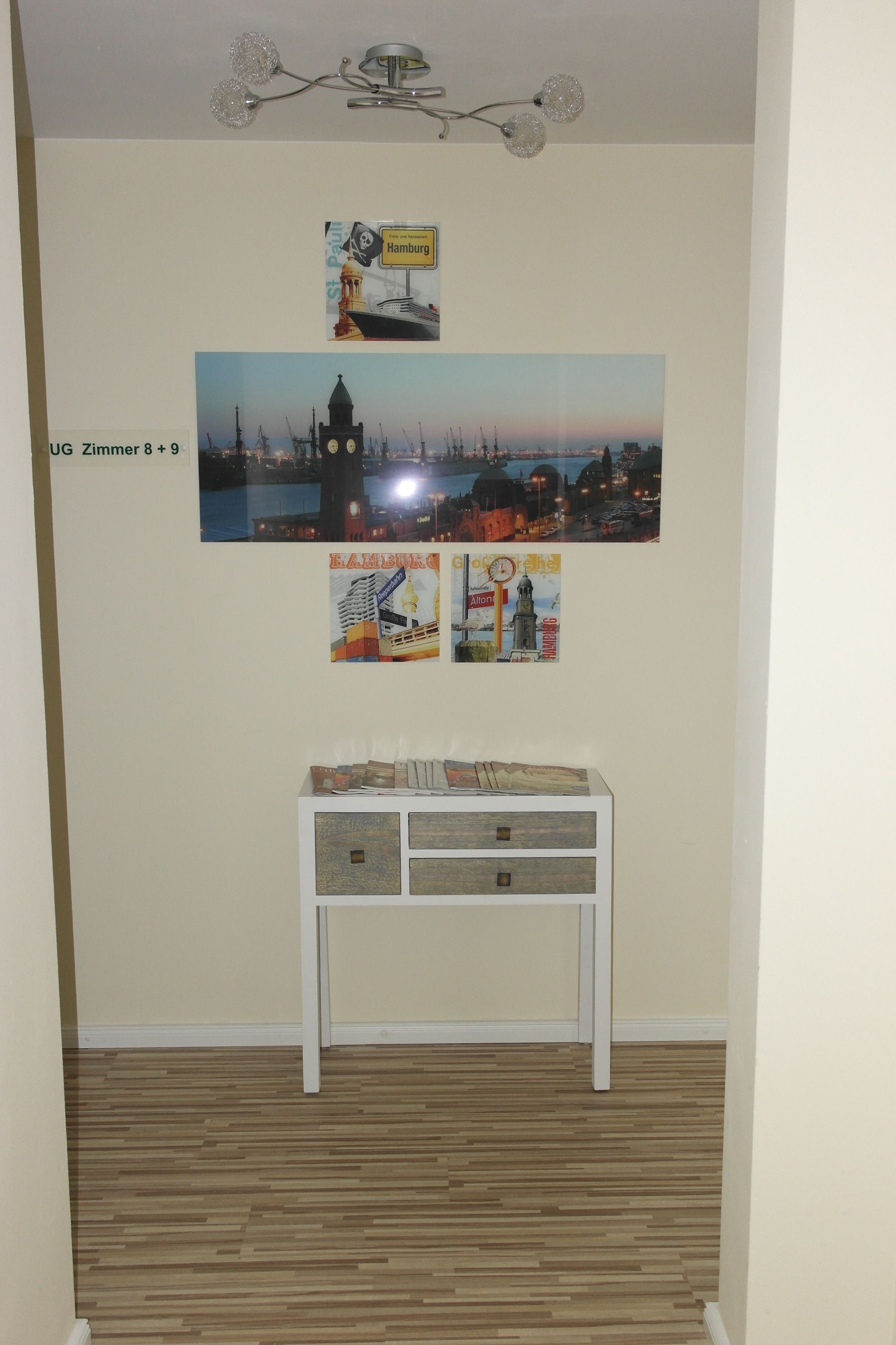 A room with a wooden floor, a white console table with drawers, and wall-mounted pictures of Hamburg and other locations.