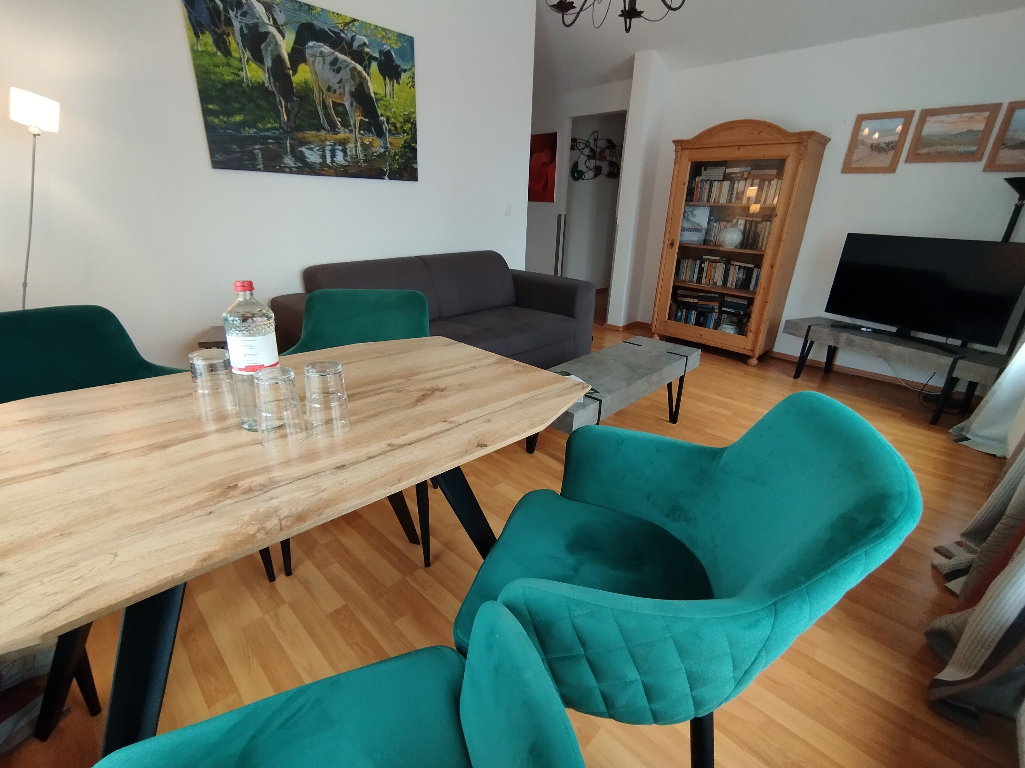A dining area with a wooden table, teal chairs, a grey sofa, a bookshelf, and a TV stand.