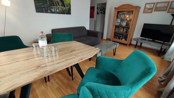 A dining area with a wooden table, teal chairs, a grey sofa, a bookshelf, and a TV stand.