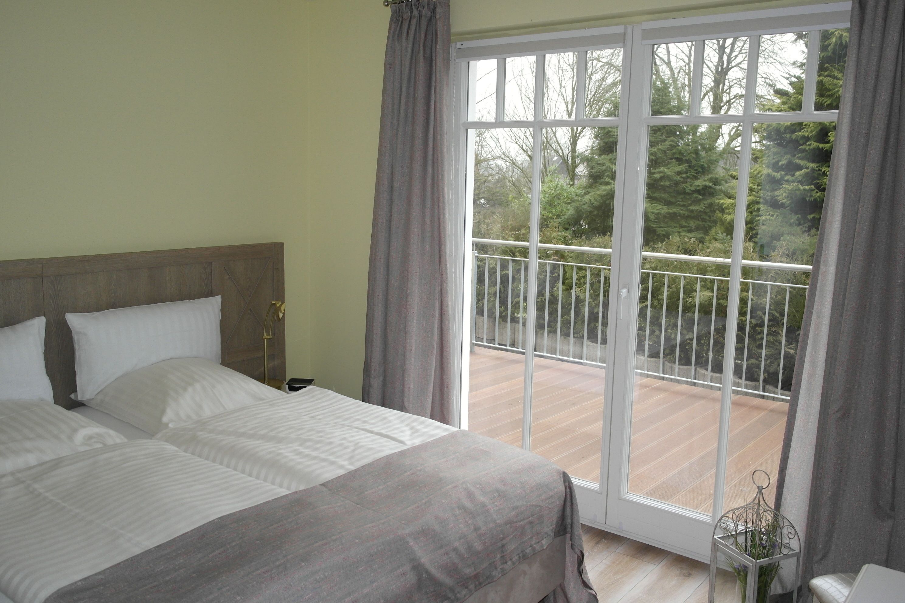 A bedroom with a large bed, a nightstand, and a window with a view of a balcony and trees.
