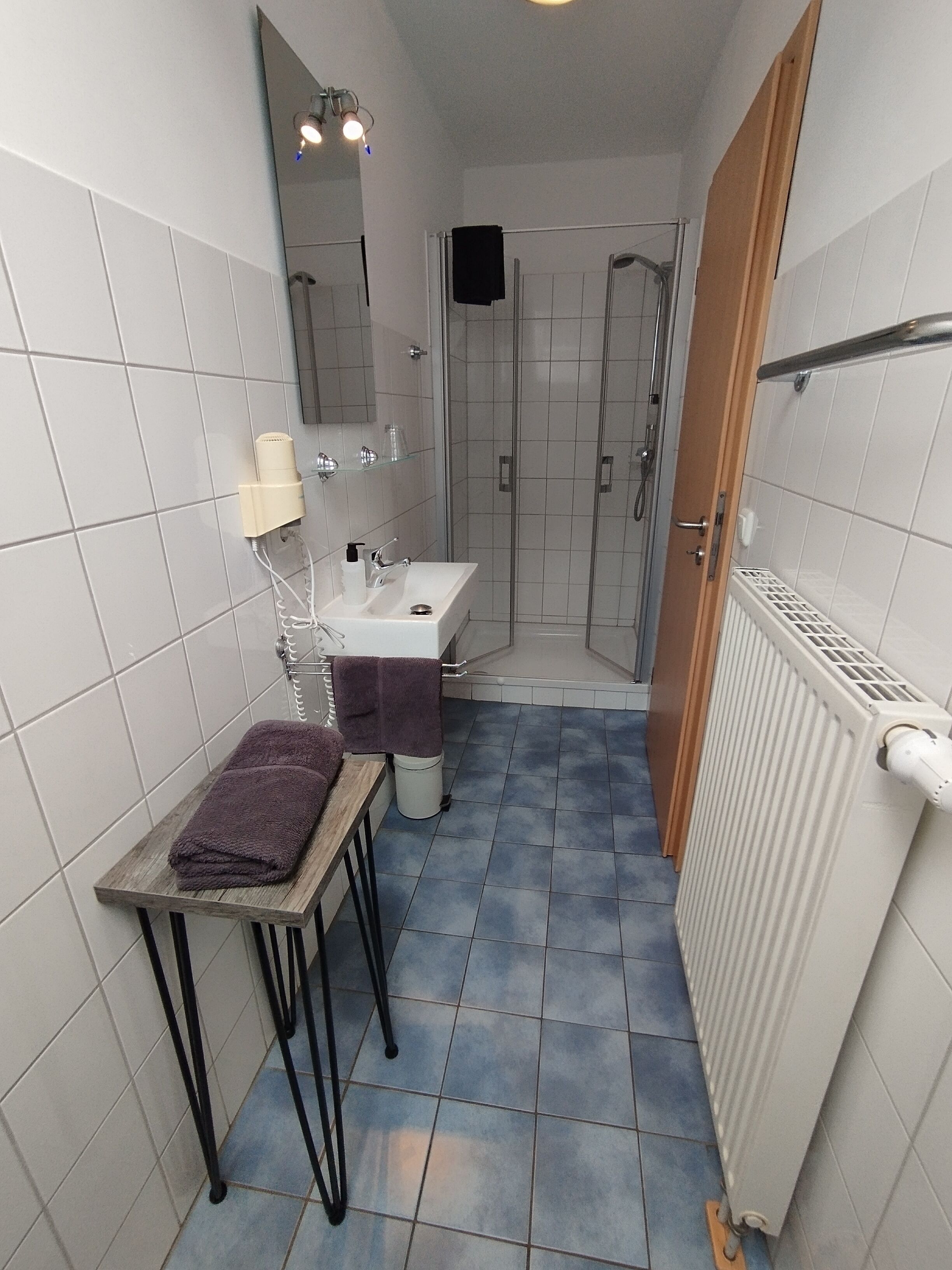 Un baño con lavamanos, espejo y ducha. Hay una toalla sobre una mesita y un radiador en la pared.