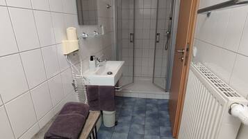 Un baño con lavamanos, espejo y ducha. Hay una toalla sobre una mesita y un radiador en la pared.