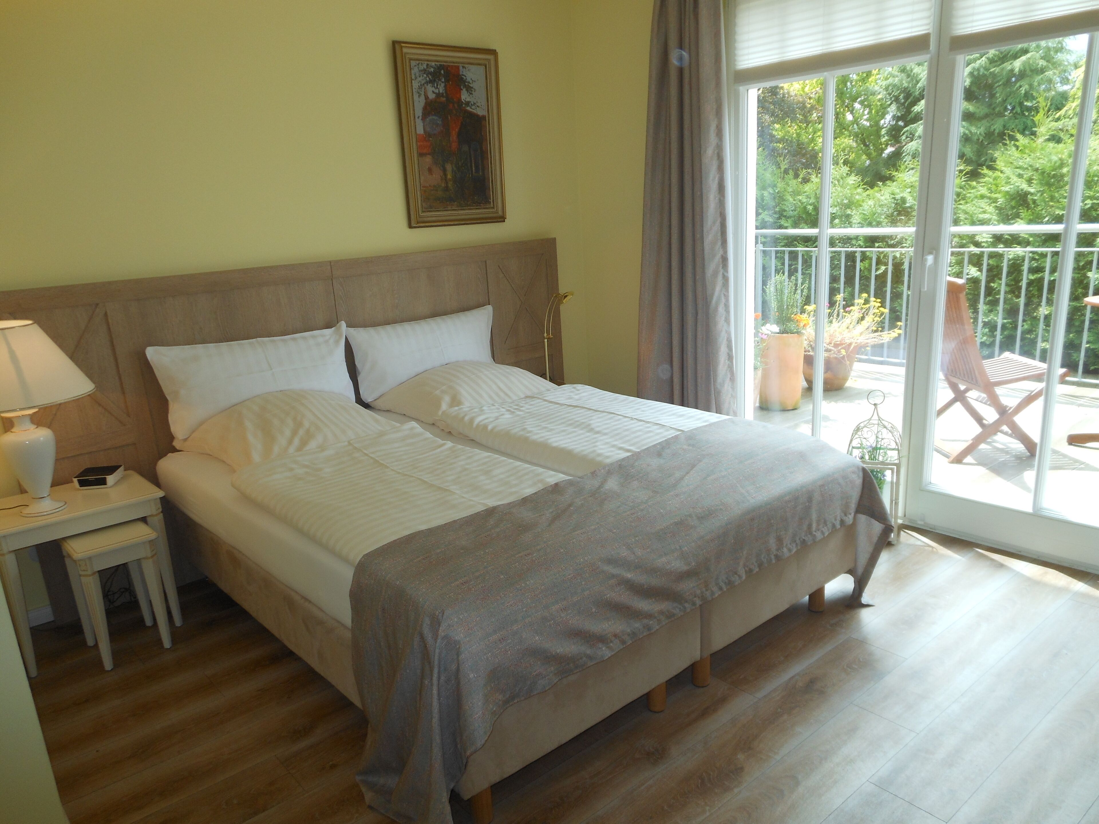 A hotel room with a large bed, a nightstand with a lamp, a painting on the wall, and a balcony with chairs and plants.