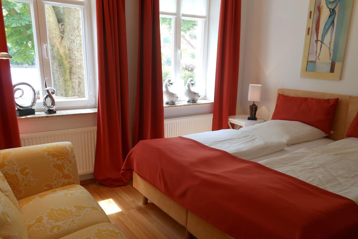Un dormitorio con cama, una silla, una ventana con cortinas, una lámpara y un cuadro en la pared.