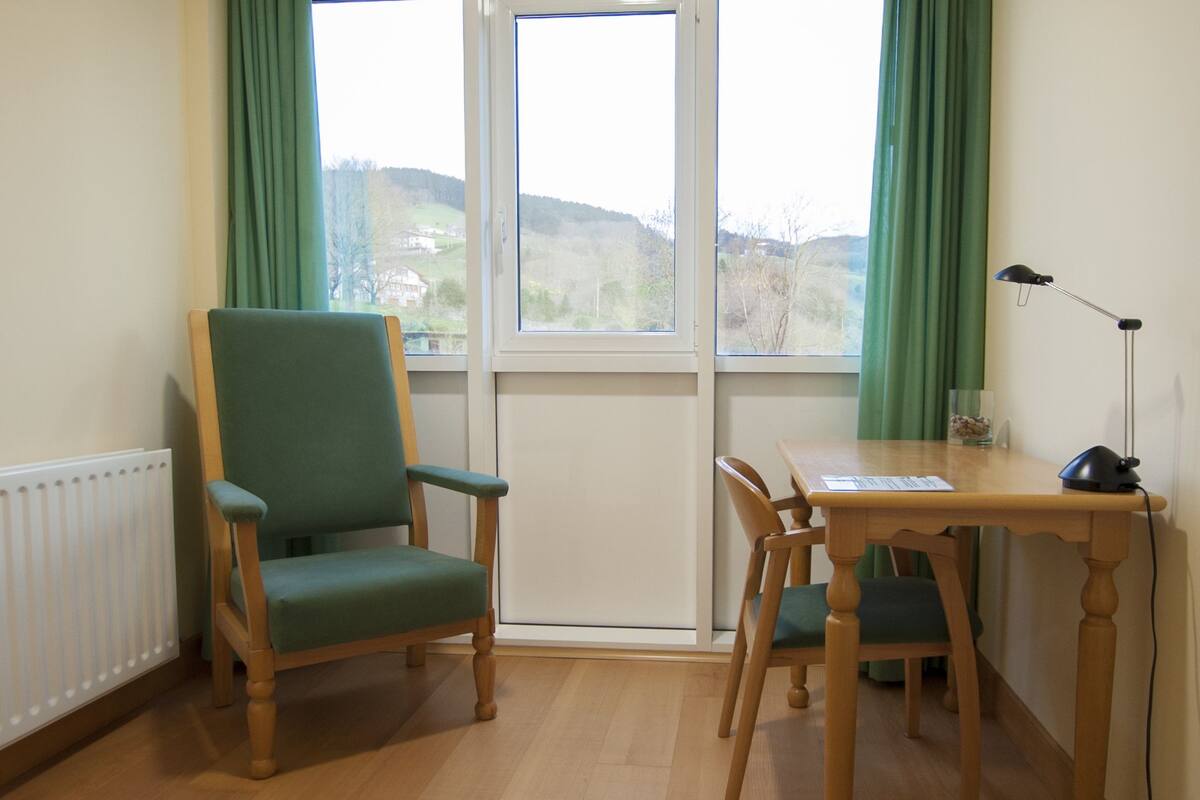 A room with a wooden chair and table, a radiator, and a view through a window.