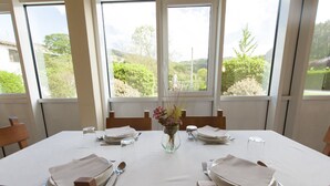 A dining table set for six with a vase of flowers, large windows, and a view of greenery. - Arrupe (Azpeitia)