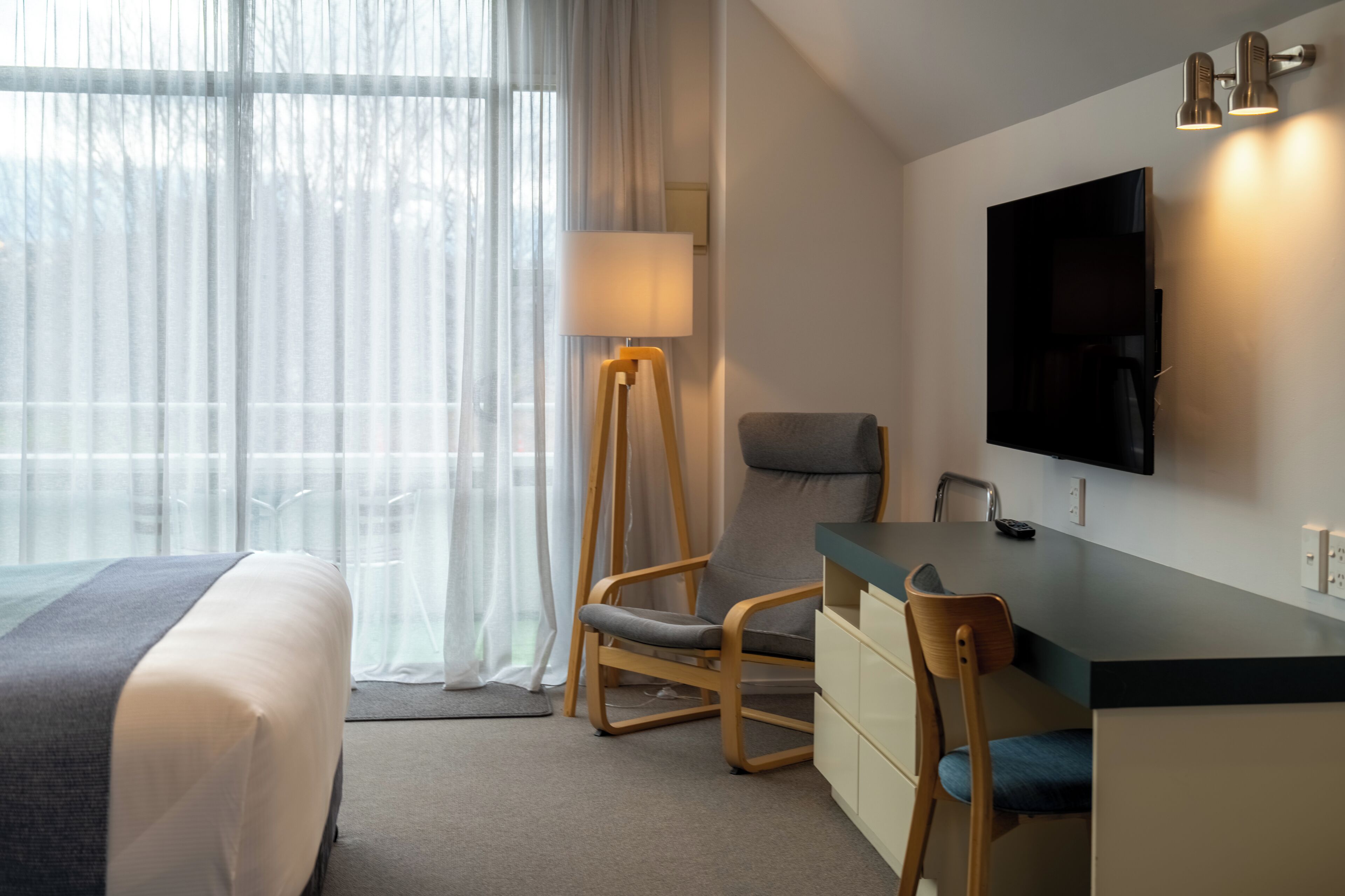 Habitación de hotel con cama, escritorio y silla, televisión montada en la pared y lámpara.