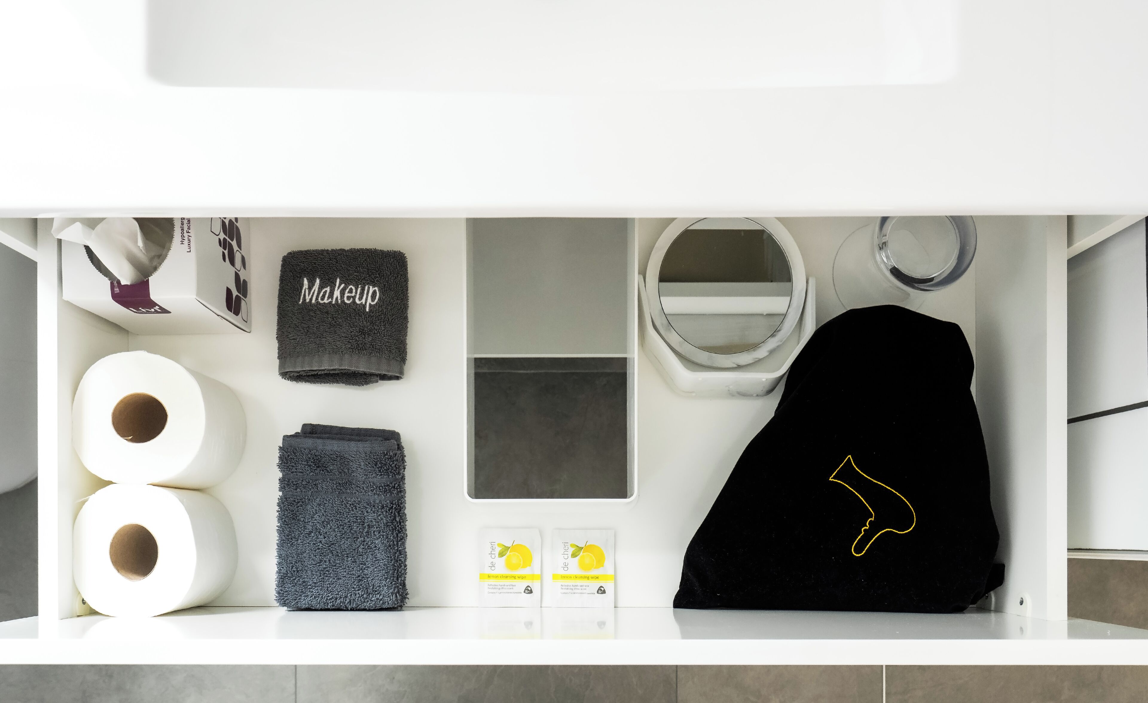 A bathroom with a white sink, a mirror, a towel rack, toilet paper, a makeup towel, a black towel, and a black bag with a yellow design.