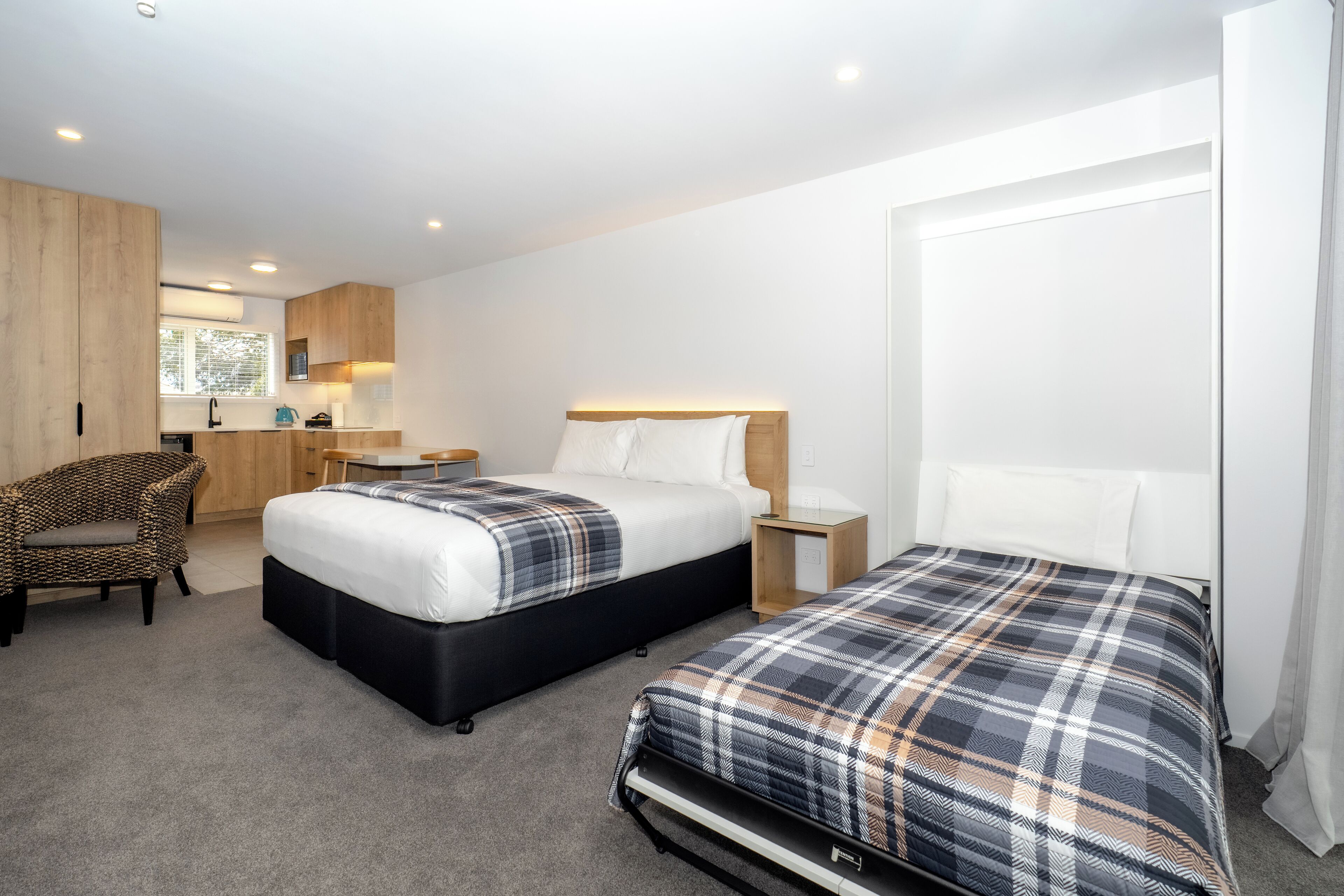 A hotel room with two beds, a kitchenette, a wooden wardrobe, and a window with a view.