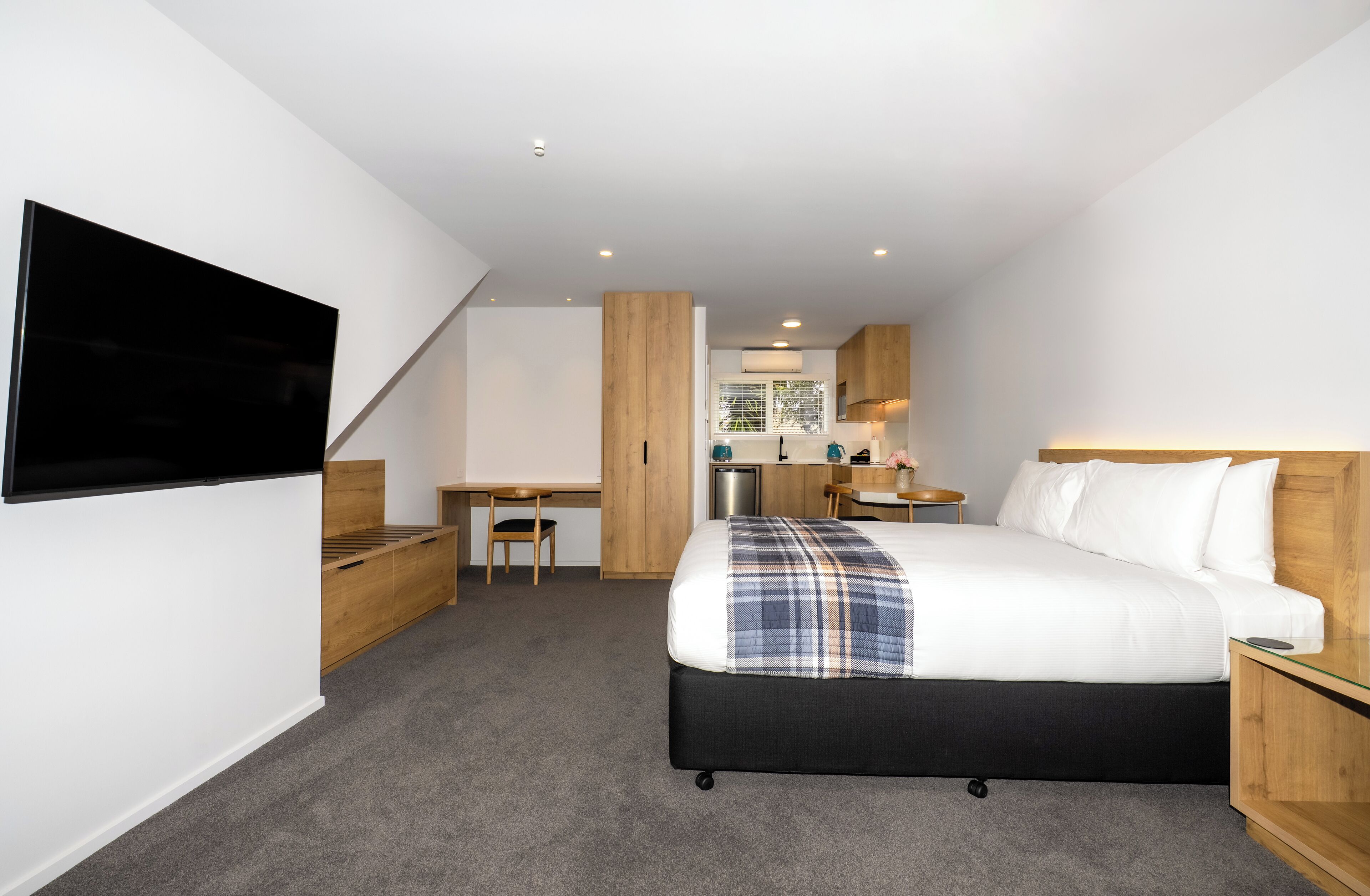 A compact hotel room with a bed, a desk, a chair, a TV, and a kitchenette.
